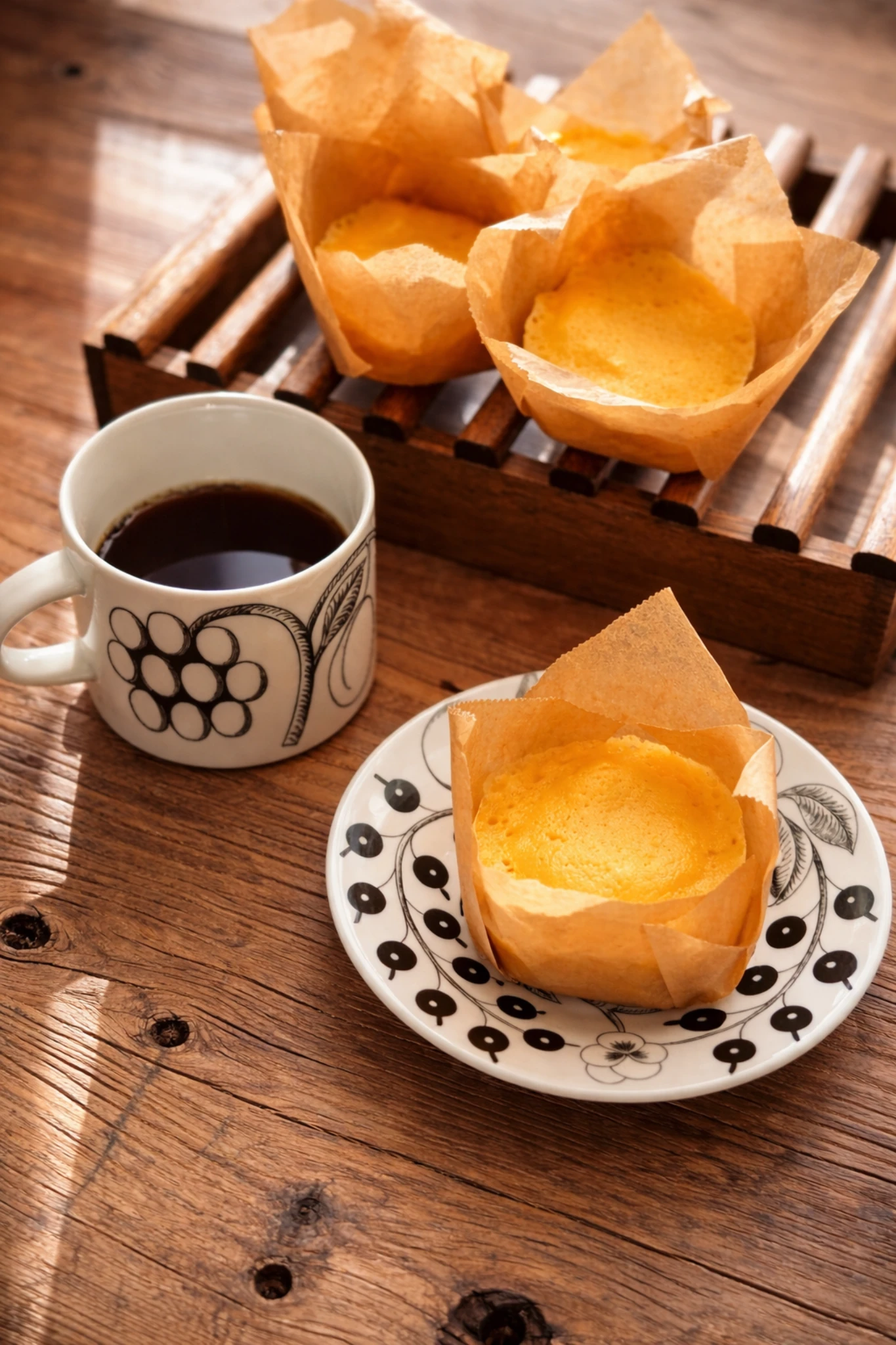 クッキングシートを敷いたカップに入ったチーズ蒸しパンとコーヒーのある木のテーブル。素朴でやさしいおやつ時間